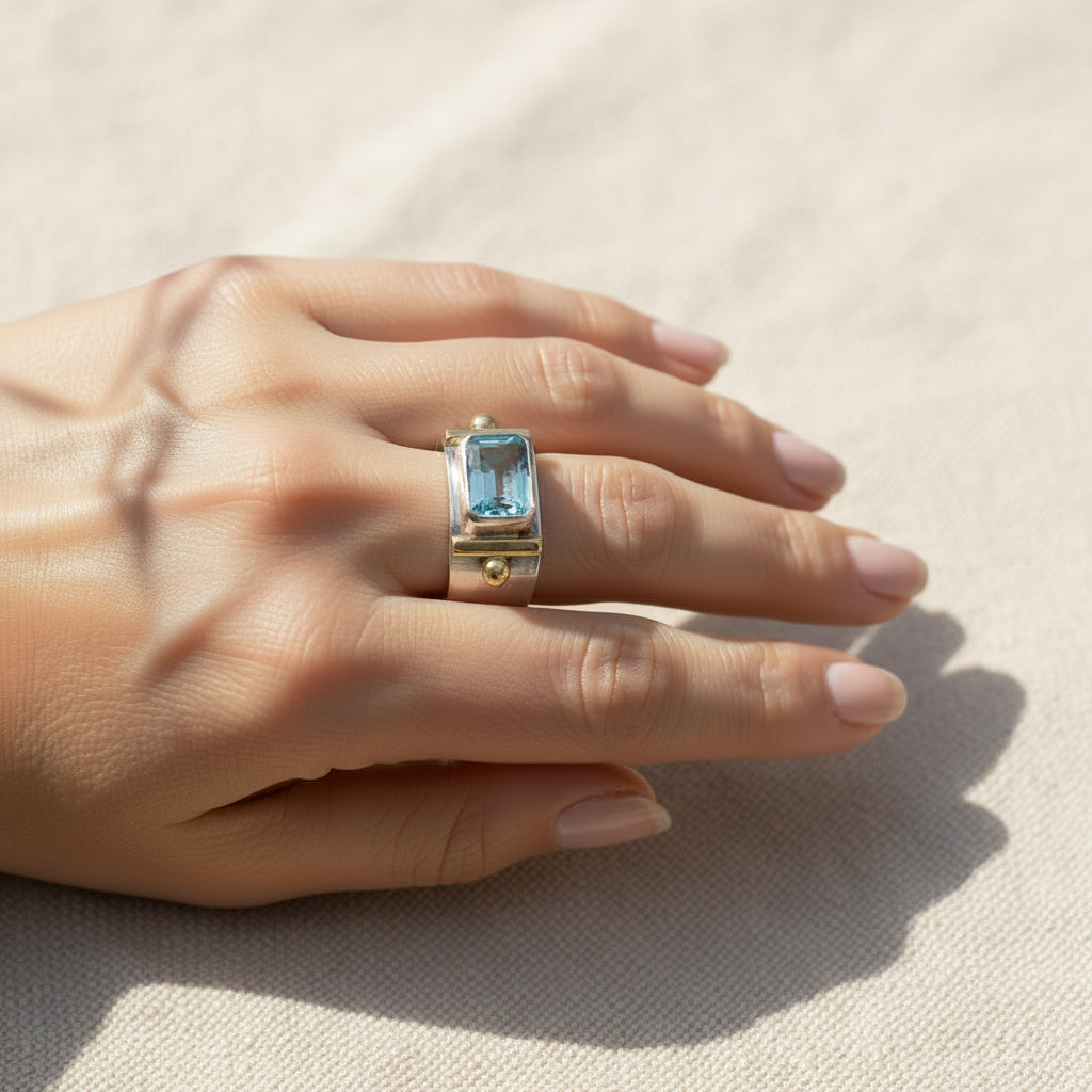 A gold ring with a rectangular light blue gemstone, worn on a person's hand against a blurred green garden background.