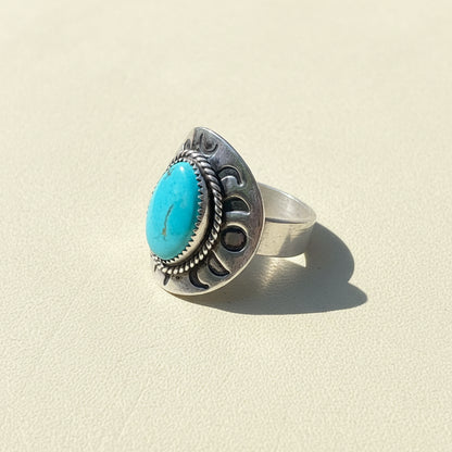 A sterling silver ring with a large oval turquoise cabochon and decorative engraved details, resting on a weathered wooden surface outdoors.
