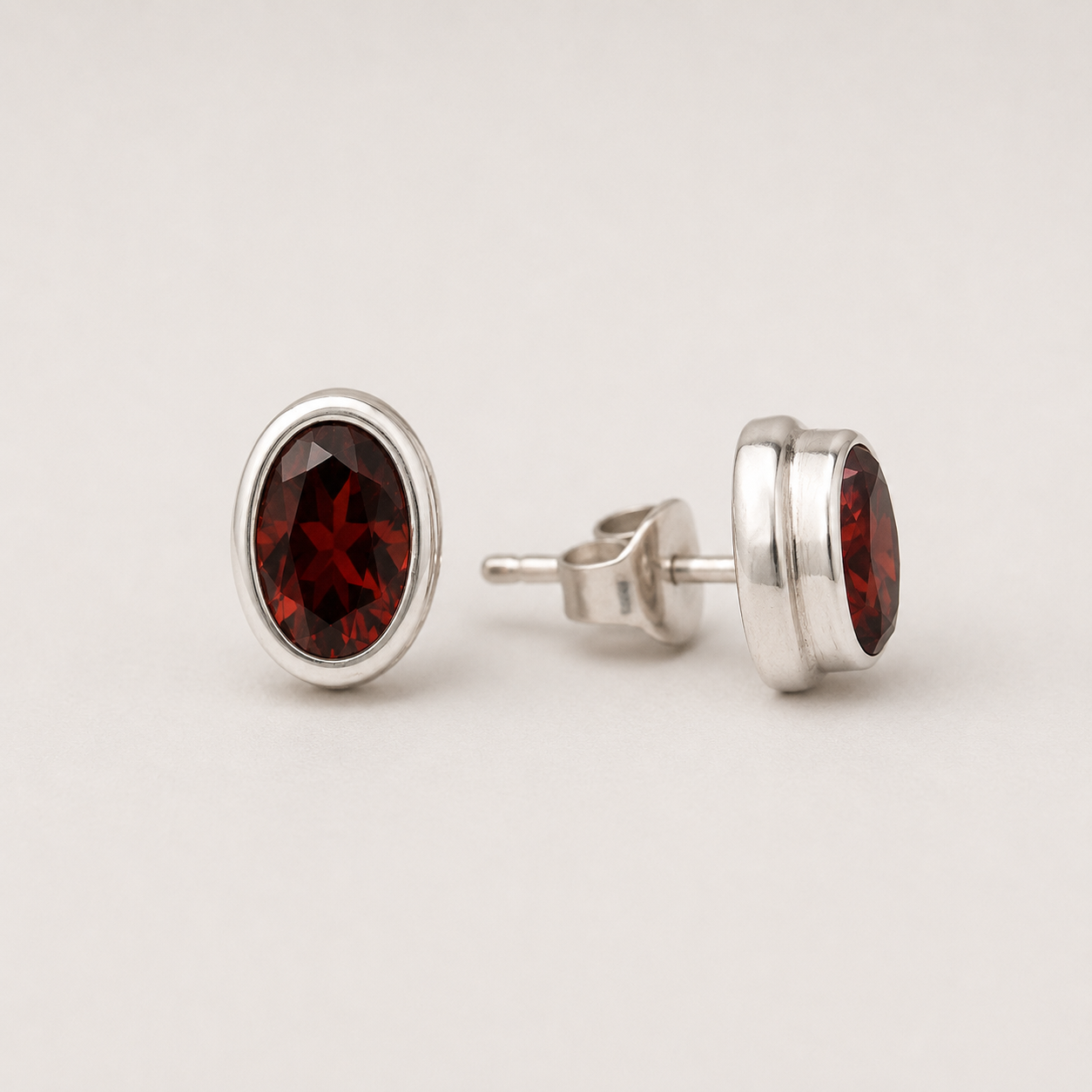 Oval garnet stud earrings with dark red faceted stone in silver bezel setting
