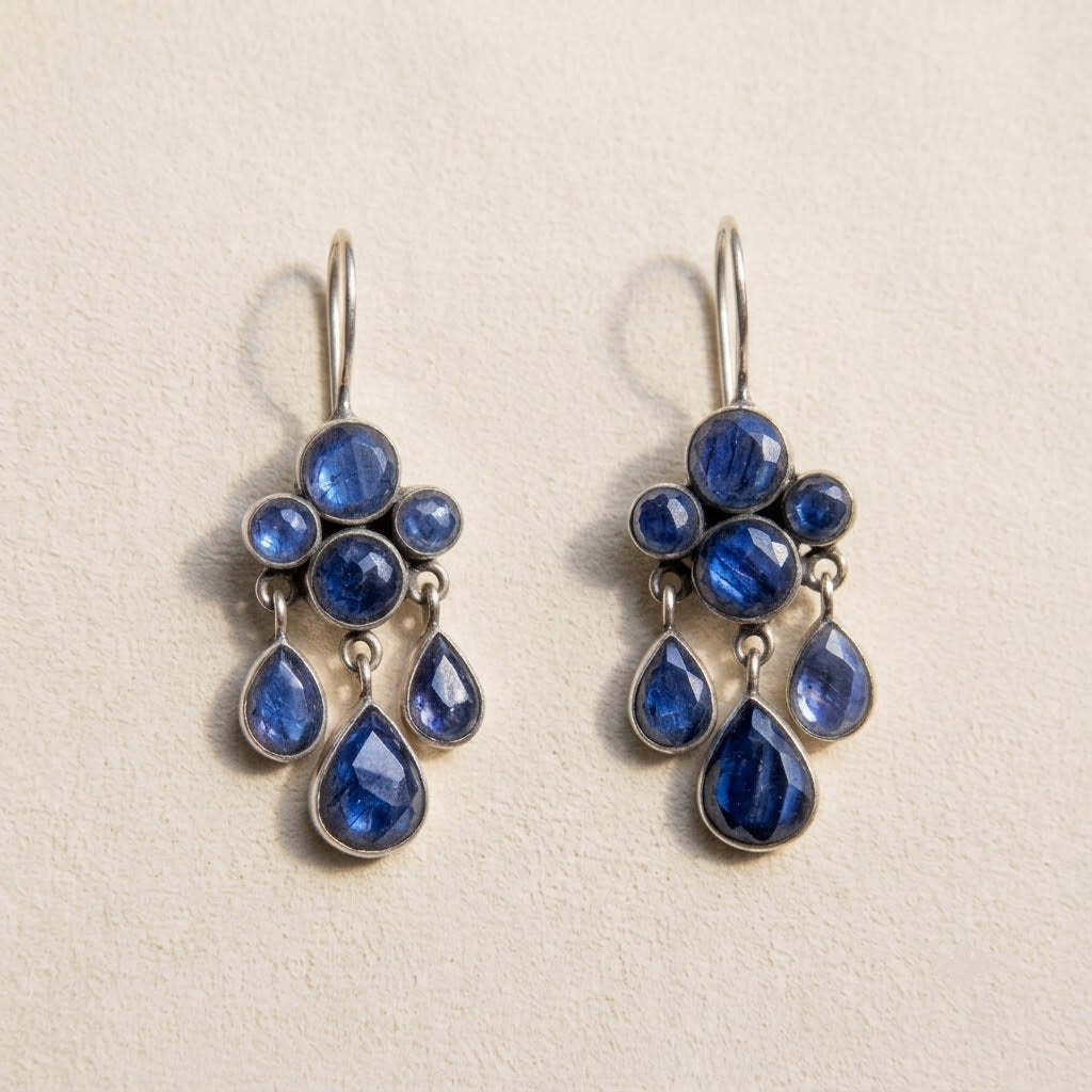 A pair of silver drop earrings featuring deep blue teardrop and round gemstones, displayed on a textured beige background.