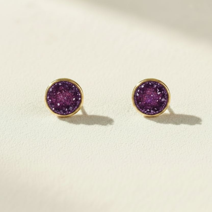 A pair of round gold stud earrings with textured purple druzy stone centers, displayed on a light cream textured surface with soft shadows.